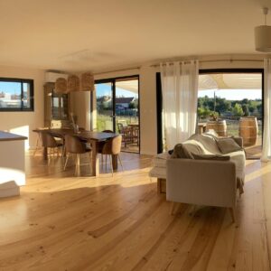 Grand espace de vie lumineux avec cuisine ouverte, salle à manger bois et salon. Terrasse avec vue sur vignoble et campagne ensoleillée.
