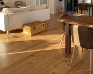 Parquet en bois clair baigné de soleil dans un salon chaleureux. Canapé blanc, malle en bois et table à manger bois naturel.