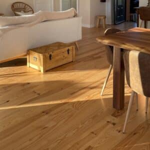 Parquet en bois clair baigné de soleil dans un salon chaleureux. Canapé blanc, malle en bois et table à manger bois naturel.